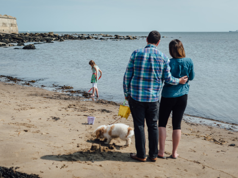 Man and wife hug on beach while watching child wade at sea edge