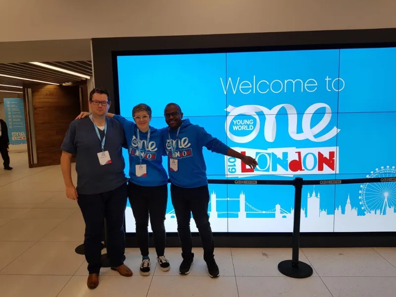 Smiling team members hug in front of One Young World London 2019 digital sign