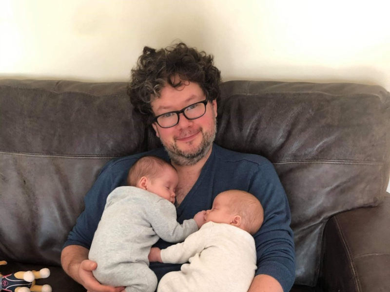 Smiling man with curly hair and glasses hodling twin babies